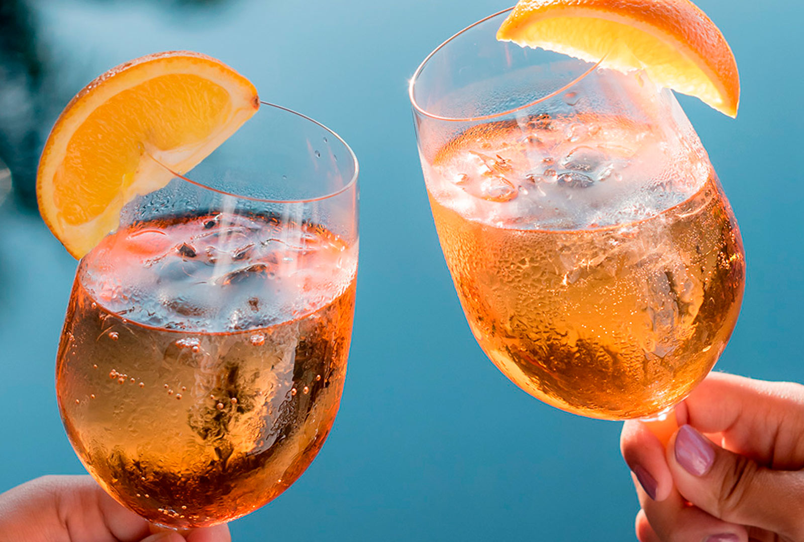 two hands holding glasses with liquid and orange slices